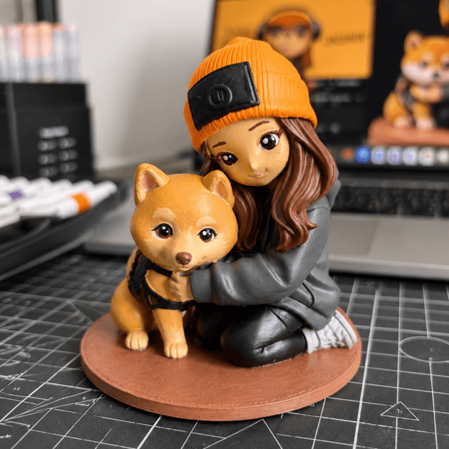 Close-up photo of a handmade Custom 3D Figure featuring a girl in an orange beanie hugging a Shiba Inu dog. The figurine sits on a round base on a crafting desk, with a blurred laptop and art supplies in the background, giving a cozy creative studio atmosphere.